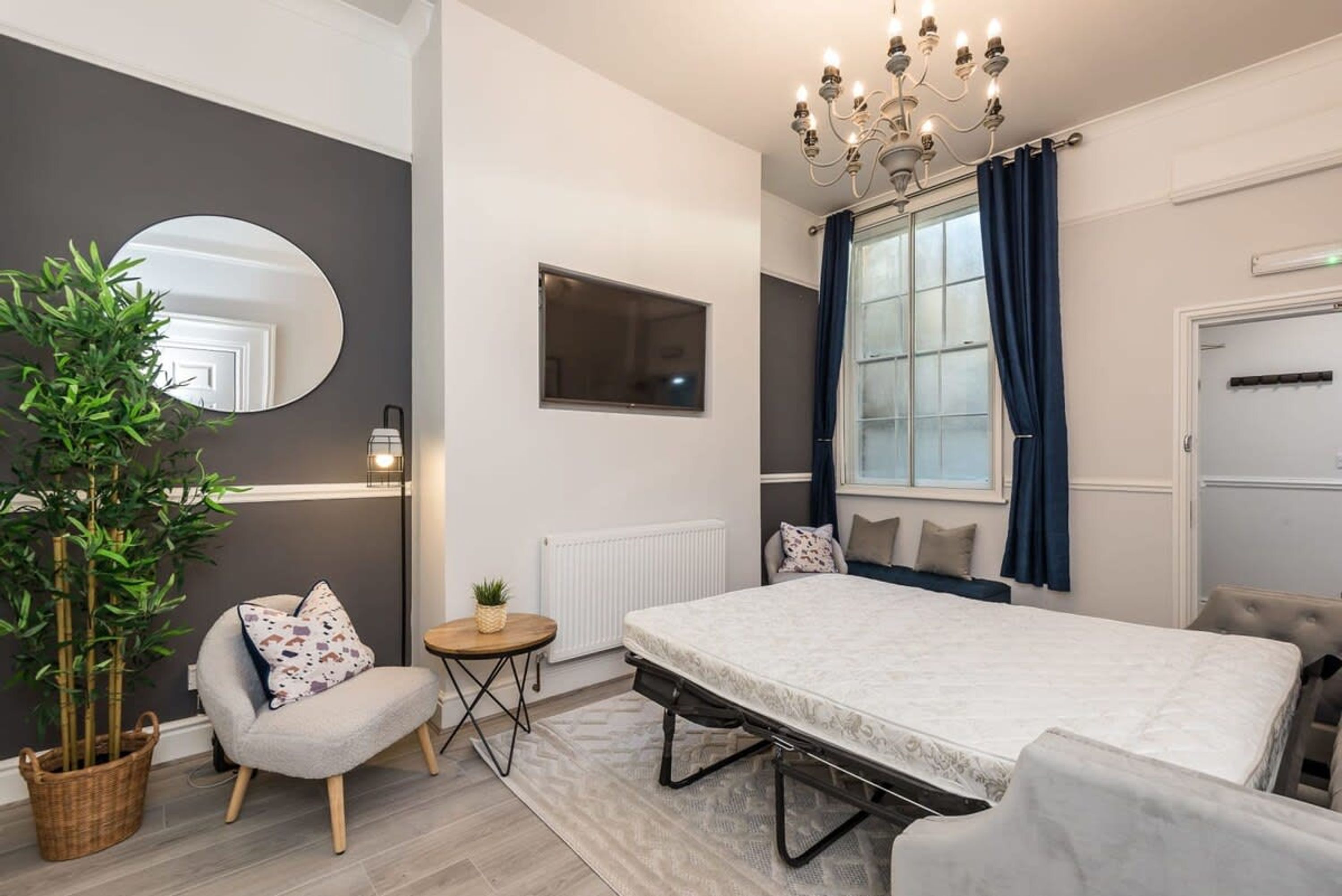 A modern bedroom with a large bed, a bedside table, a chair, a potted plant, a mirror, and a window with curtains.