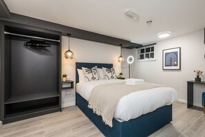 Ein modernes Schlafzimmer mit einem großen Bett, einem dunklen Wandschrank, Pendelleuchten, einem Nachttisch und einem kleinen Fenster.