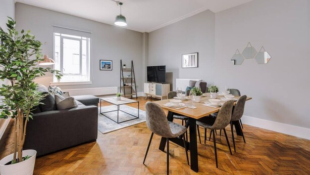 A modern living room with a dining area, a grey sofa, a coffee table, and a potted plant. - Host Apartments The Bank Apt (Liverpool)