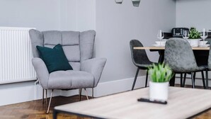 A modern living room with a grey armchair, a dining table with chairs, and a kitchen area in the background. - Host Apartments The Bank Apt (Liverpool)