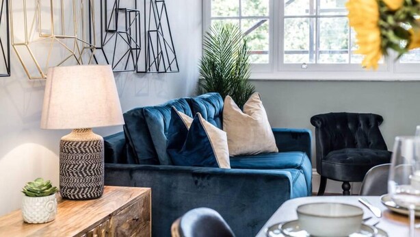 A modern living room with a blue sofa, a black chair, a wooden side table with a lamp, and a potted plant.
