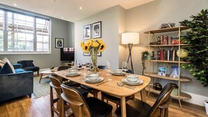 A dining area with a wooden table set for four, surrounded by chairs. There is a bookshelf with books and decorative items, a large window providing natural light, and a sofa in the background.