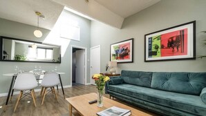 En moderne stue med en grøn sofa, et træbord med en vase blomster, samt et spiseområde med et glasbord og stole.