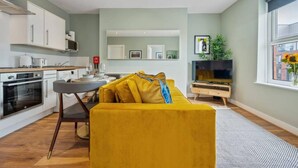 A modern kitchen with white cabinets, a dining area with a yellow sofa, and a large window with a view of the city. - Host Apartments The Georgian Quarters at Bedford (Liverpool)