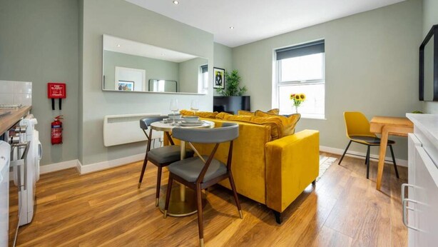 A modern living room with a yellow sofa, a dining table with chairs, a large mirror, and a window with a view of the outdoors. - Host Apartments The Georgian Quarters at Bedford (Liverpool)