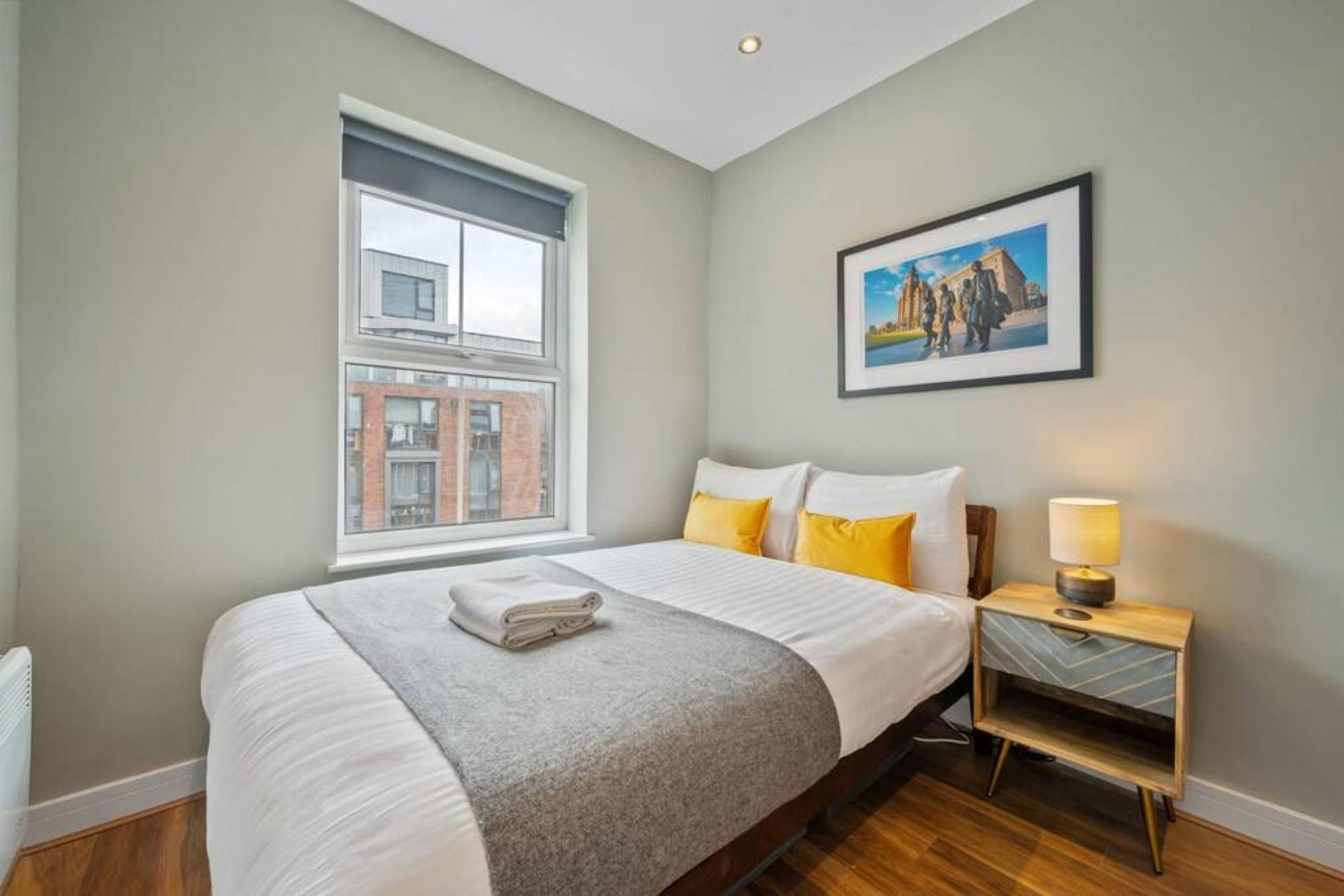 A hotel room with a large bed, a window with a view of buildings, a framed picture on the wall, and a bedside table with a lamp.