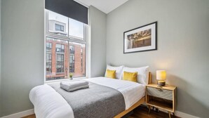 A modern hotel room with a large bed, a bedside table with a lamp, a framed picture on the wall, and a window with a view of buildings. - Host Apartments The Georgian Quarters at Bedford (Liverpool)