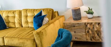 A yellow sofa with blue cushions, a wooden side table with a lamp, and a geometric wall decoration.