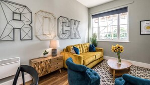 A living room with a yellow sofa, blue armchairs, a wooden side table, and a lamp. There are abstract wall art pieces and a window with a view. - Host Stay Catharine Quarters II (Liverpool)