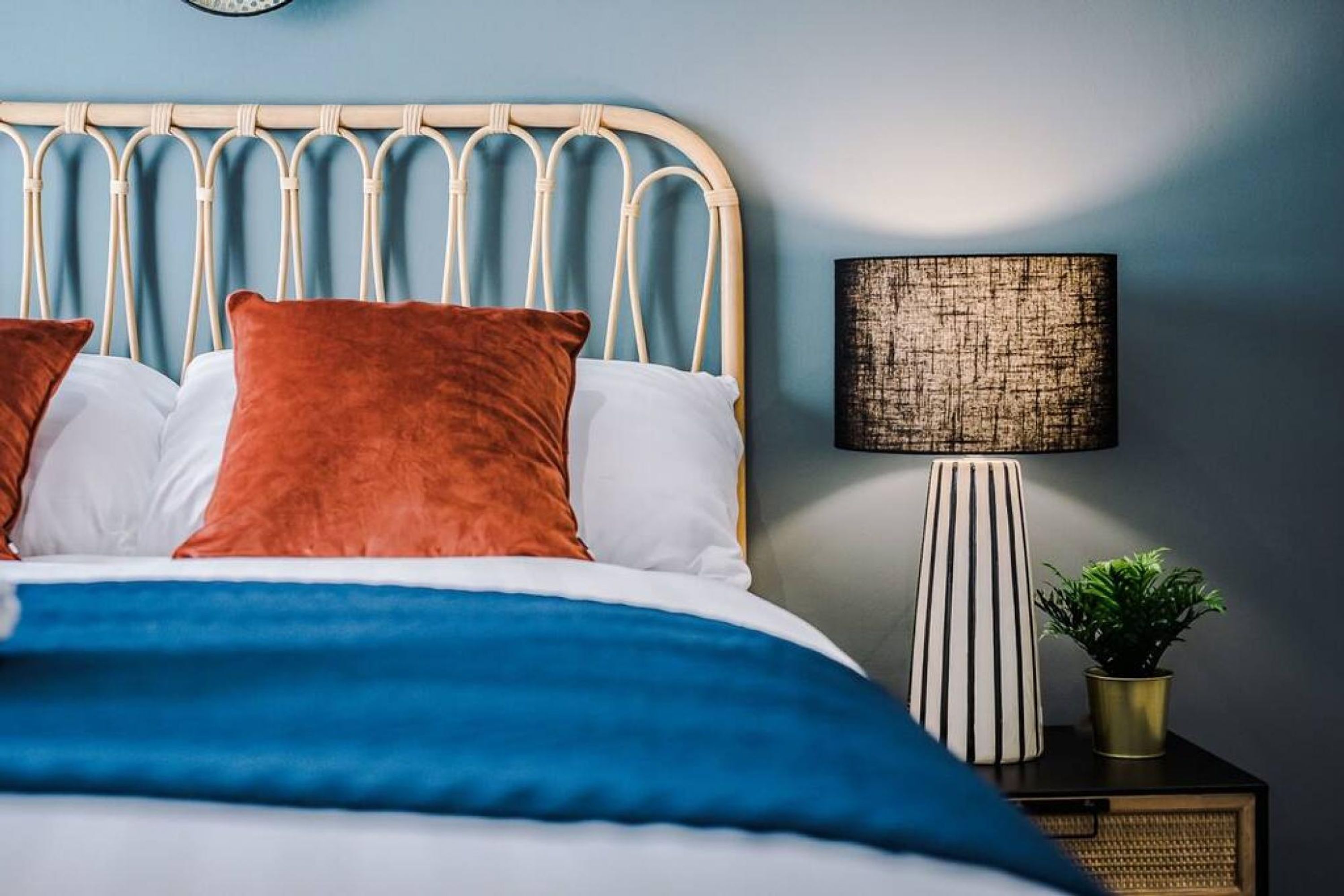 A bed with a blue and white duvet, two orange pillows, and a white pillow. There is a lamp with a striped shade and a small plant on a bedside table.