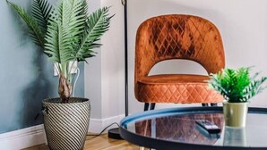A modern living room with a round glass coffee table, a brown upholstered armchair, a tall floor lamp, and a large potted plant.