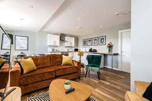 A modern living room with a brown sofa, a wooden coffee table, and a green armchair. A kitchen with white cabinets and a sink is visible in the background. - Host Apartments Catharine Quarters III (Liverpool)