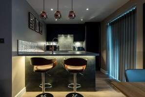 A modern kitchen with a central island, two bar stools, pendant lighting, and a view of the living area with a window and blinds. - Host Apartments The Pilgrim Coach House II (Liverpool)
