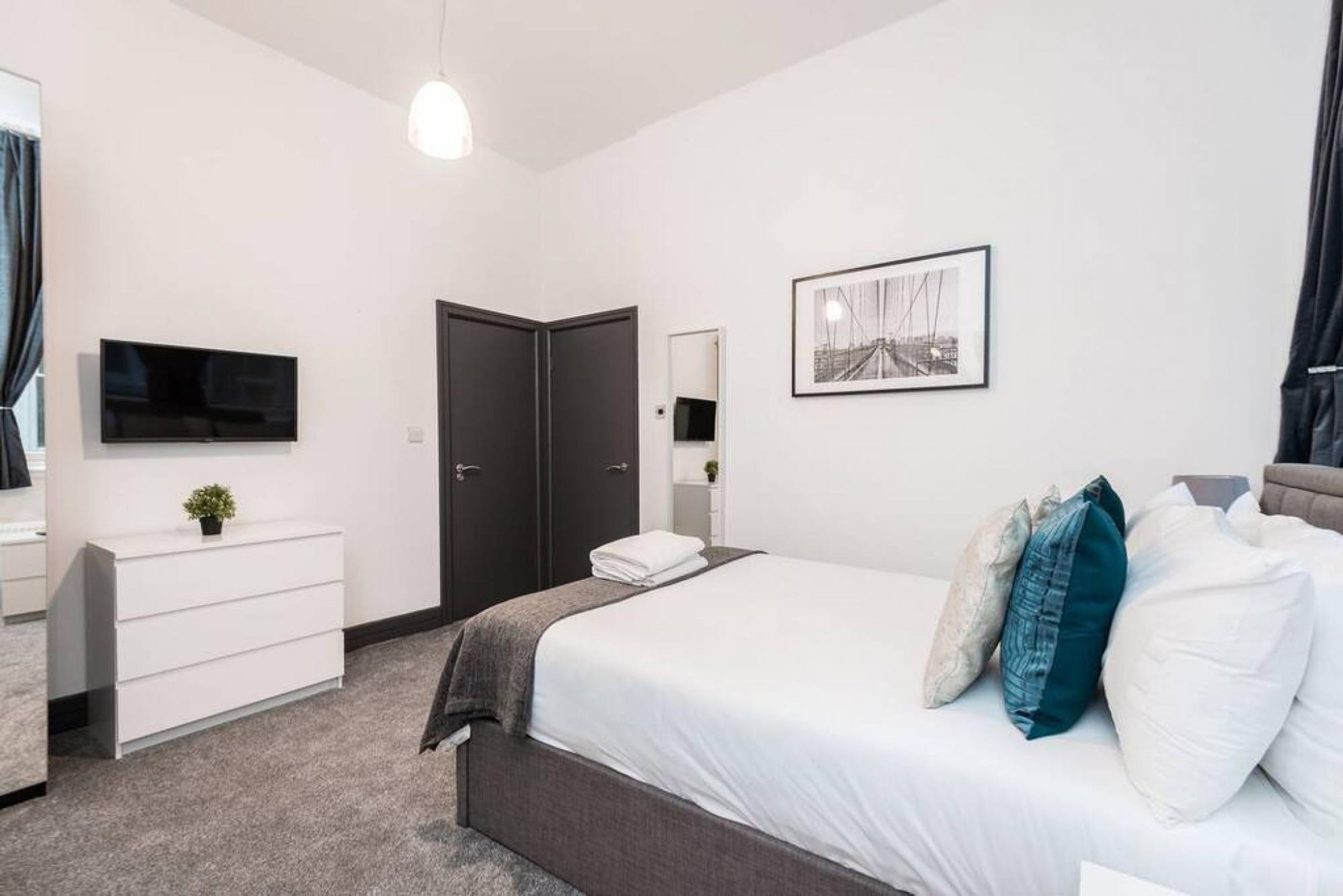 Un dormitorio moderno con cama, cómoda, televisor, puerta y un cuadro en la pared.