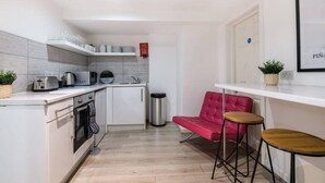 Une cuisine de taille réduite avec des meubles blancs, une poubelle en acier inoxydable, une chaise rouge et deux tabourets.