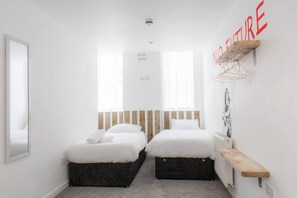 A minimalist bedroom with two beds, a wooden headboard, a small shelf, and a wall-mounted rack.