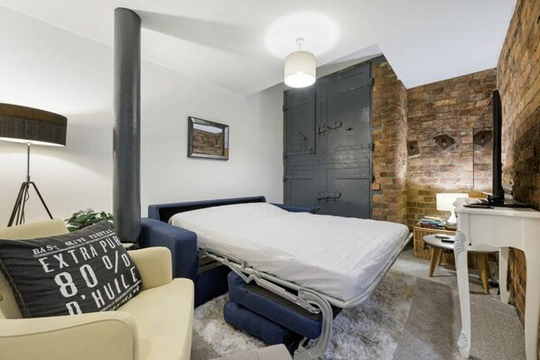 A bedroom with a bed, a chair, a desk, a lamp, and a brick wall. - Host Apartments Henry s Hideaway (Liverpool)