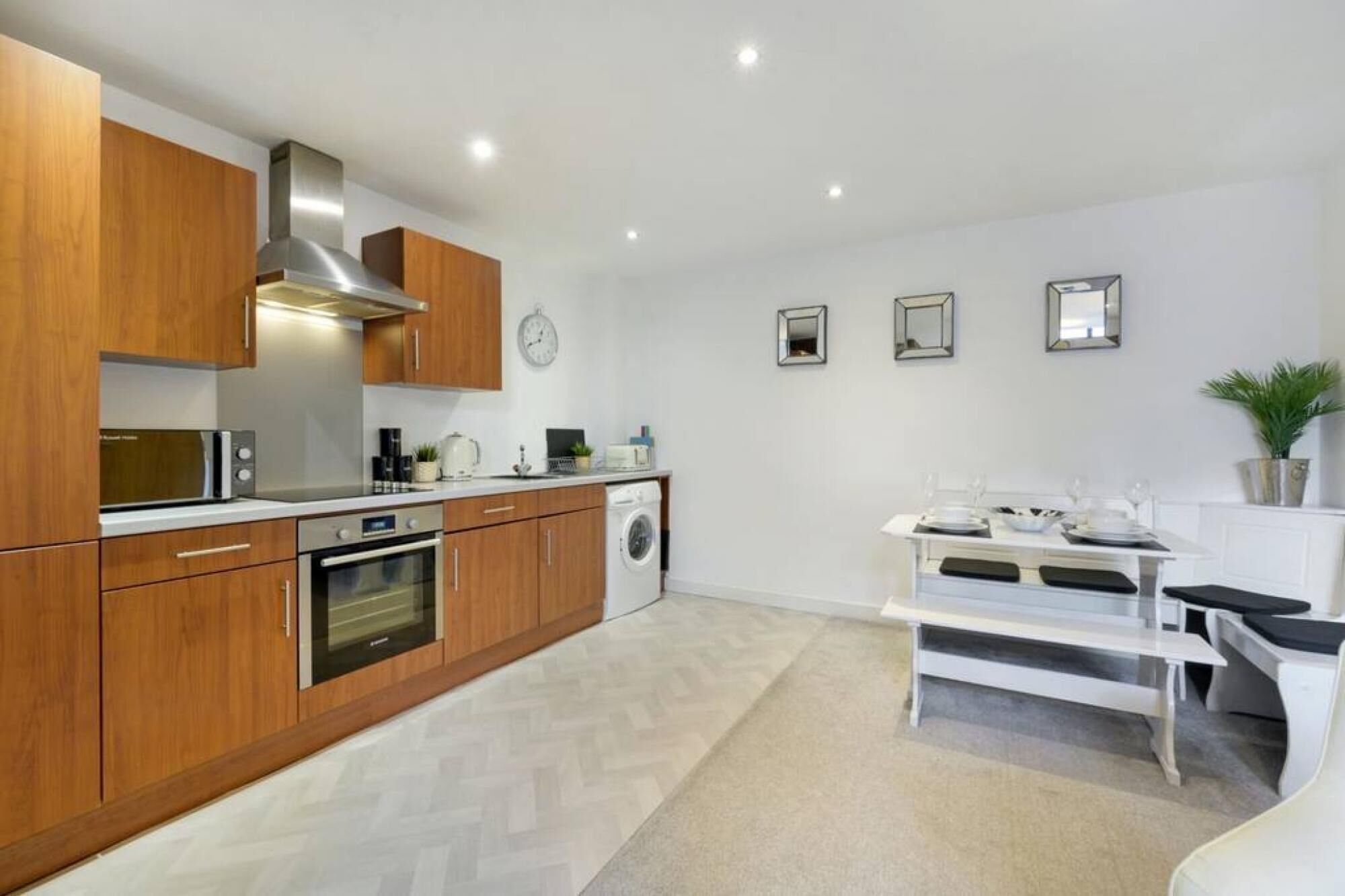 A modern kitchen with wooden cabinets, a built-in oven, a microwave, and a washing machine. There is a dining table with chairs and a potted plant on the right.