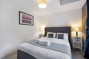 A neatly made bed with white and grey linens, two bedside tables with lamps, a framed picture on the wall, and a window with a curtain. - Host Apartments Henry s Hideaway (Liverpool)