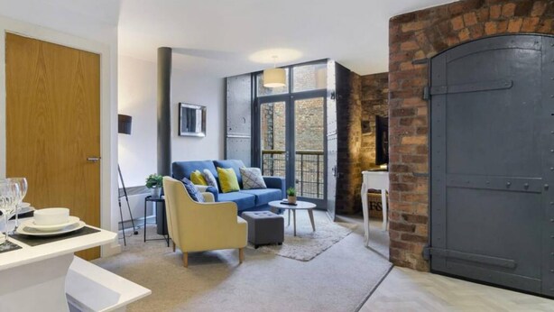 A modern living room with a blue sofa, a yellow armchair, and a small round table. There is a large wooden door and a brick wall on one side.