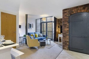 A modern living room with a blue sofa, a yellow armchair, and a small round table. There is a large wooden door and a brick wall on one side. - Host Apartments Henry s Hideaway (Liverpool)