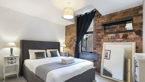 A modern bedroom with a large bed, a brick wall, a window with curtains, a mirror, and a bedside table with a lamp.