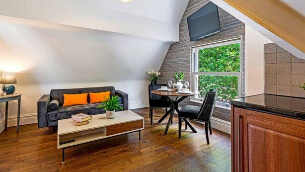 Un espace de vie moderne de style loft, comprenant un coin repas, un canapé avec des coussins orange, une table basse et une grande fenêtre donnant sur un paysage verdoyant.