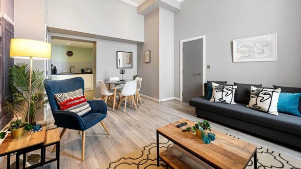 A modern living room with a blue armchair, a dark sofa, and a coffee table. There is a dining area with a table and chairs, and a kitchen in the background. - Host Apartments Halifax House II (Liverpool)