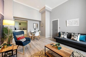 Een moderne woonkamer met een blauwe fauteuil, een donkere bank en een salontafel. Er is een eethoek met een tafel en stoelen, en een keuken op de achtergrond.