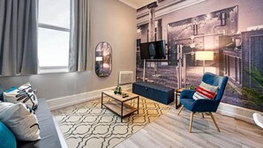 A modern living room with a large window, a flat-screen TV, a blue armchair, and a unique wall mural. - Host Apartments Halifax House II (Liverpool)