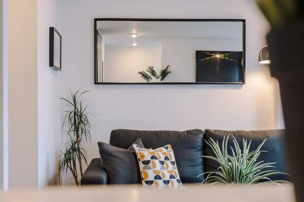 Ein modernes Wohnzimmer mit einem dunkelgrauen Sofa, einem großen Spiegel und zwei Zimmerpflanzen.