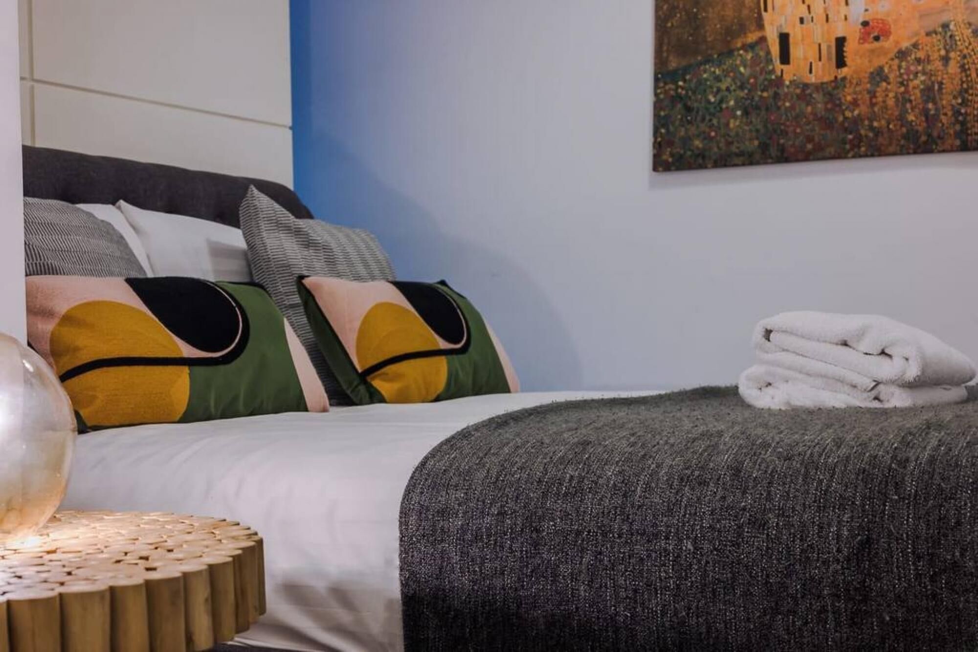 A neatly made bed with a grey blanket and pillows, a white towel on the side, and a painting on the wall.