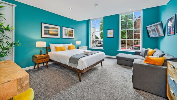 A bedroom with teal walls, a large bed, a grey sofa, and a window with a view of the street. - Host Apartments The Georgian Quarters at Canning (Liverpool)