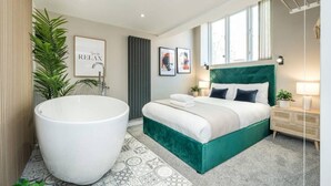 Ein modernes Schlafzimmer mit einer großen Badewanne, einem grün gepolsterten Bett und Blick auf die Stadt durch große Fenster.