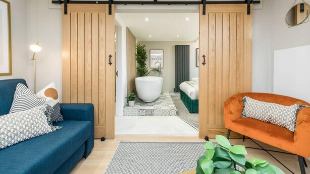 Ein modernes Wohnzimmer mit einem blauen Sofa, einem orangefarbenen Sessel und einer großen Holztür, die sich zur Badezimmer öffnet.