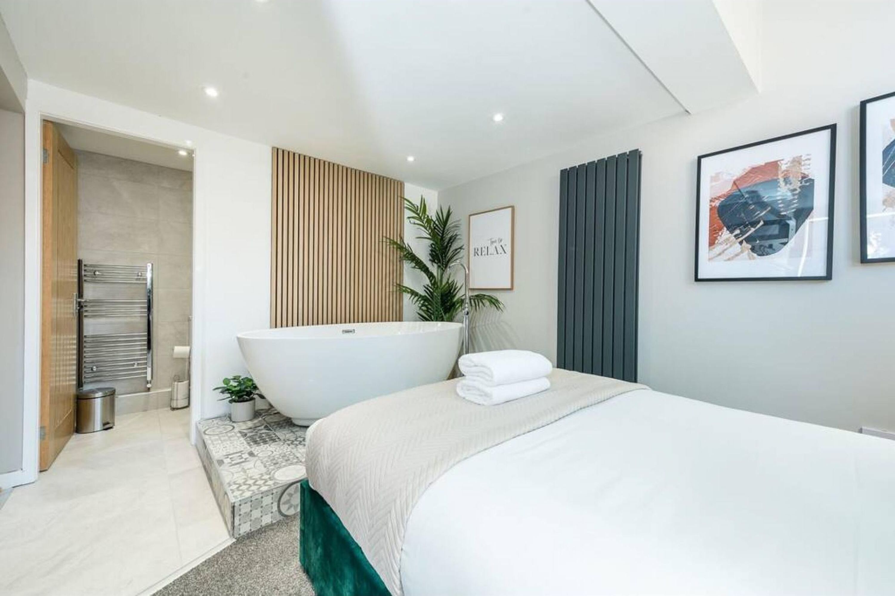 Quarto moderno com cama grande, banheira independente, uma pequena planta em vaso e uma obra de arte emoldurada na parede.