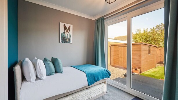 A bedroom with a bed, pillows, a blue blanket, a wall art of a dog, and a sliding glass door leading to a garden. - Host Stay Sands Walk Cottage (Redcar)