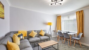 A modern living room with a grey sectional sofa, yellow cushions, a wooden coffee table, and a dining area with a table and chairs. - Host Stay Sands Walk Cottage (Redcar)