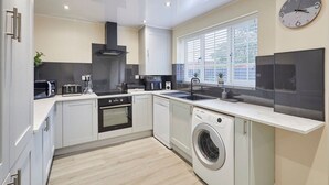 A modern kitchen with white cabinets, a built-in oven, a microwave, a washing machine, and a large window with blinds. - Host Stay Violet s View (Whitby)