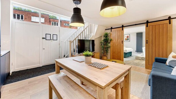 A modern kitchen with a wooden table, a bench, and pendant lighting. There is a staircase leading to a loft area, and a sliding door to a bedroom. - Host Apartments The Georgian Coach House 2 (Liverpool)