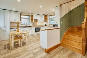 A modern kitchen with white cabinetry, a wooden dining table, and a wooden staircase. - Host Stay Fawcett Cottage (York)