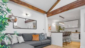 Uma sala de estar moderna com um sofá cinzento, vigas de madeira, uma mesa de centro e uma cozinha ao fundo.