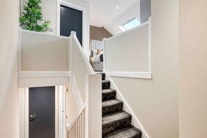 A staircase with a dark carpeted handrail and white walls, leading to a room with a bed and a window.
