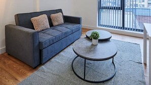 Un salon moderne avec un canapé bleu foncé, deux œuvres d’art encadrées, une petite table ronde ornée d’une plante et une grande fenêtre avec des stores.