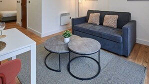 Un salon moderne avec un canapé gris, deux tables rondes, une petite plante en pot et un tableau abstrait encadré accroché au mur.