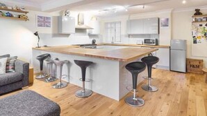 A modern kitchen with a central island, bar stools, a grey sofa, and a wooden floor. - Host Stay Cobb Court (Margate)