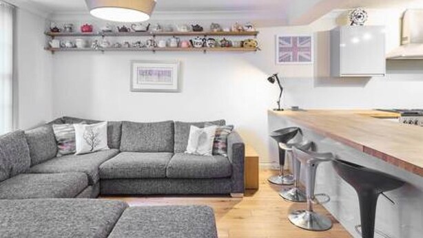 A modern living room with a grey sectional sofa, a wooden coffee table, and a kitchen area with a bar stool and a white countertop. - Host Stay Cobb Court (Margate)
