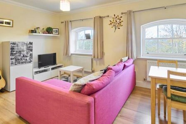 A living room with a pink sofa, a TV, a small table, and a window with curtains. - Host Stay Nunnery Fields (Canterbury)
