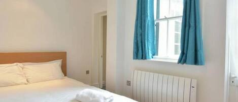 A bedroom with a wooden bed, white bedding, a window with blue curtains, and a white radiator.