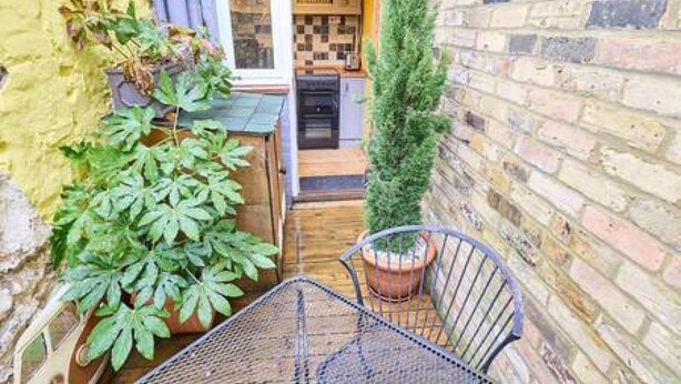 Un espacio exterior estrecho con una mesa y sillas de metal, una planta en maceta y un muro de ladrillo.